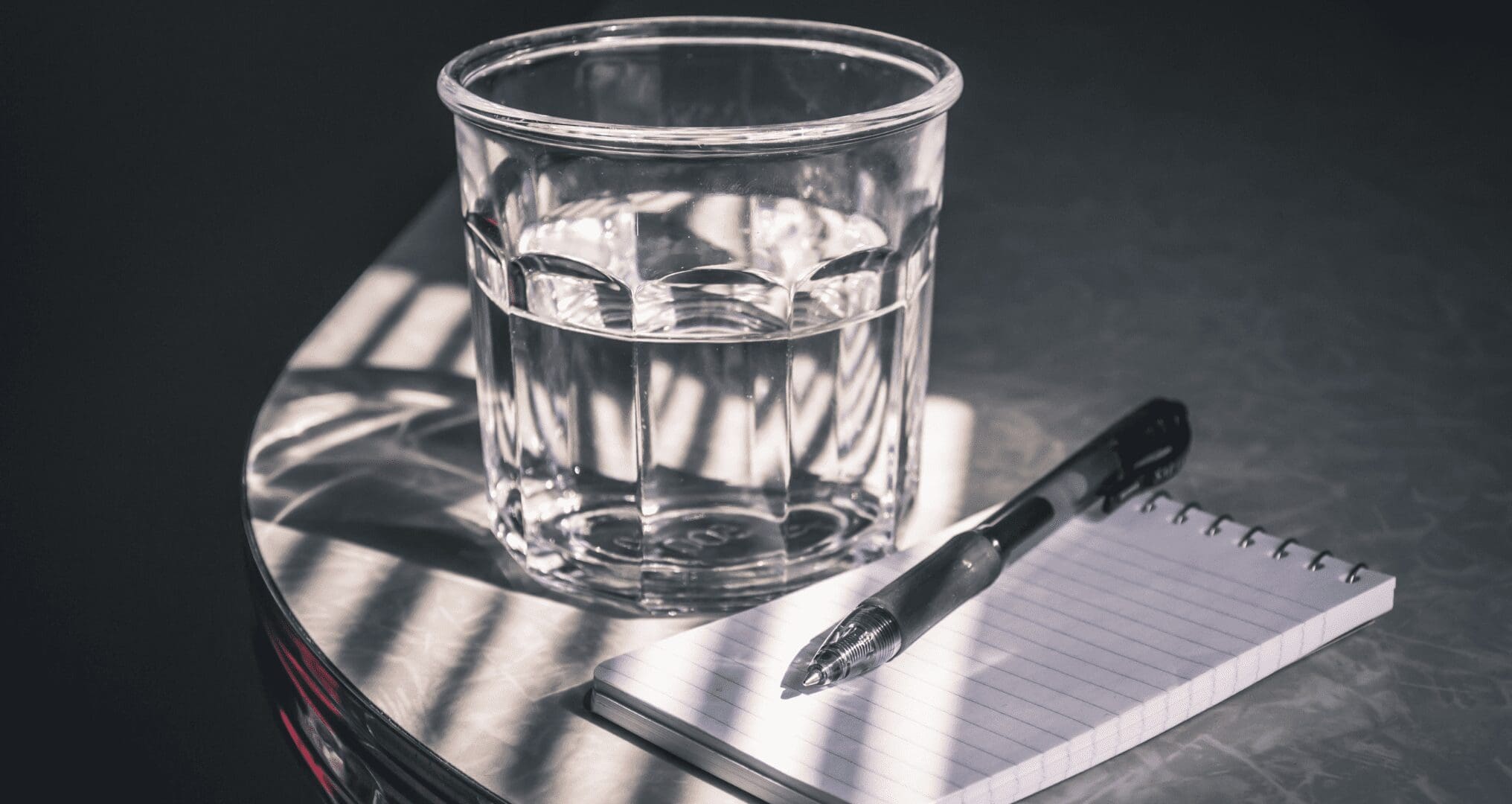 _A glass of water beside a pen and a notepad A glass of water beside a pen and a notepad.